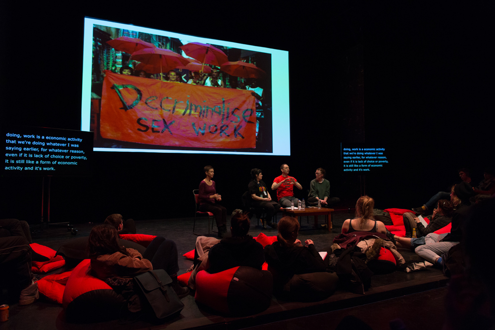 A projected image reads "decriminalise sex work" on a red banner. Four people are sat underneath the image on a stage talking with an audience.