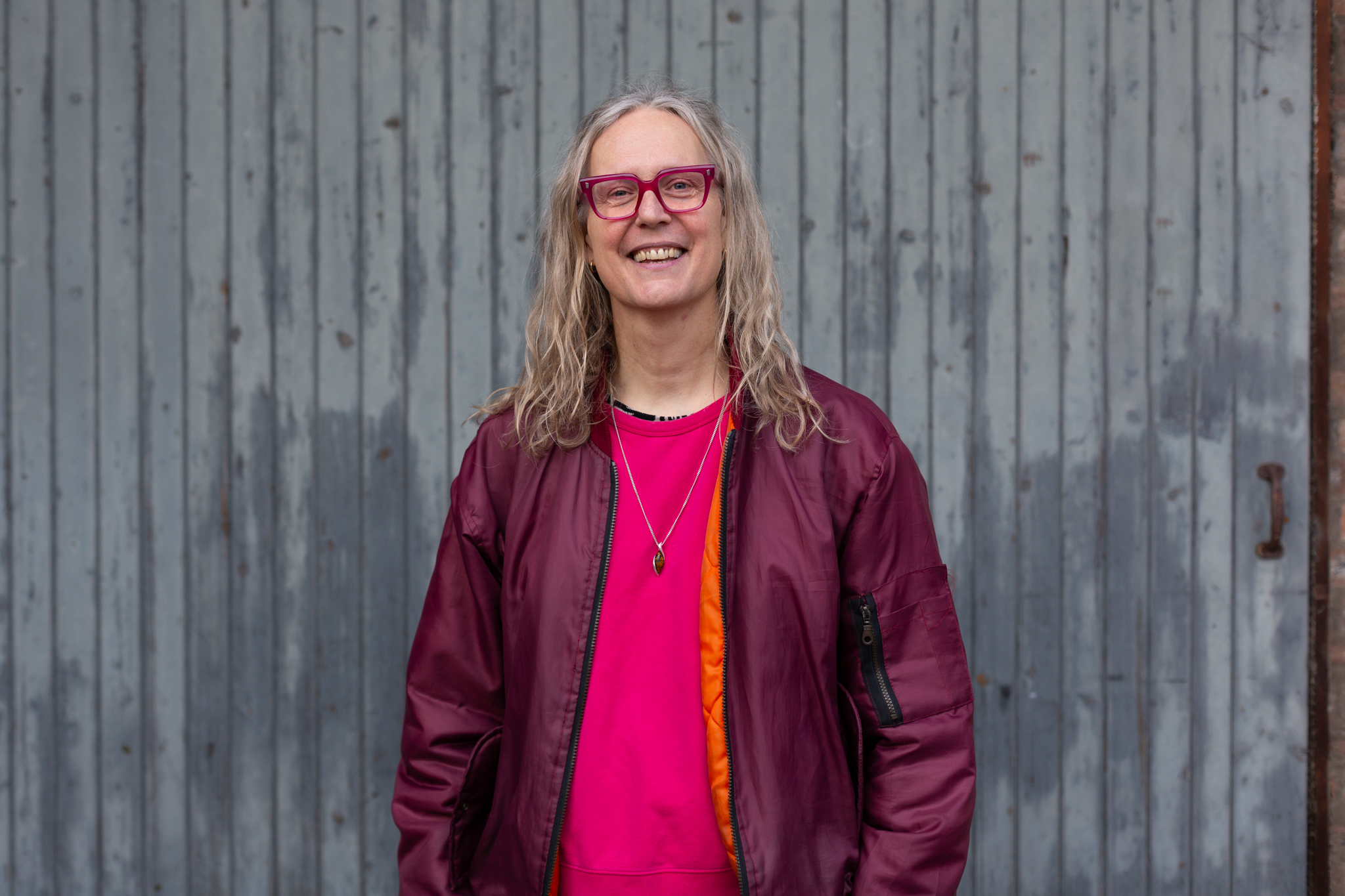 Mijke van der Drift smiles, wearing bright pink rimmed glasses, a bright pink jumper under an unzipped short maroon jacket