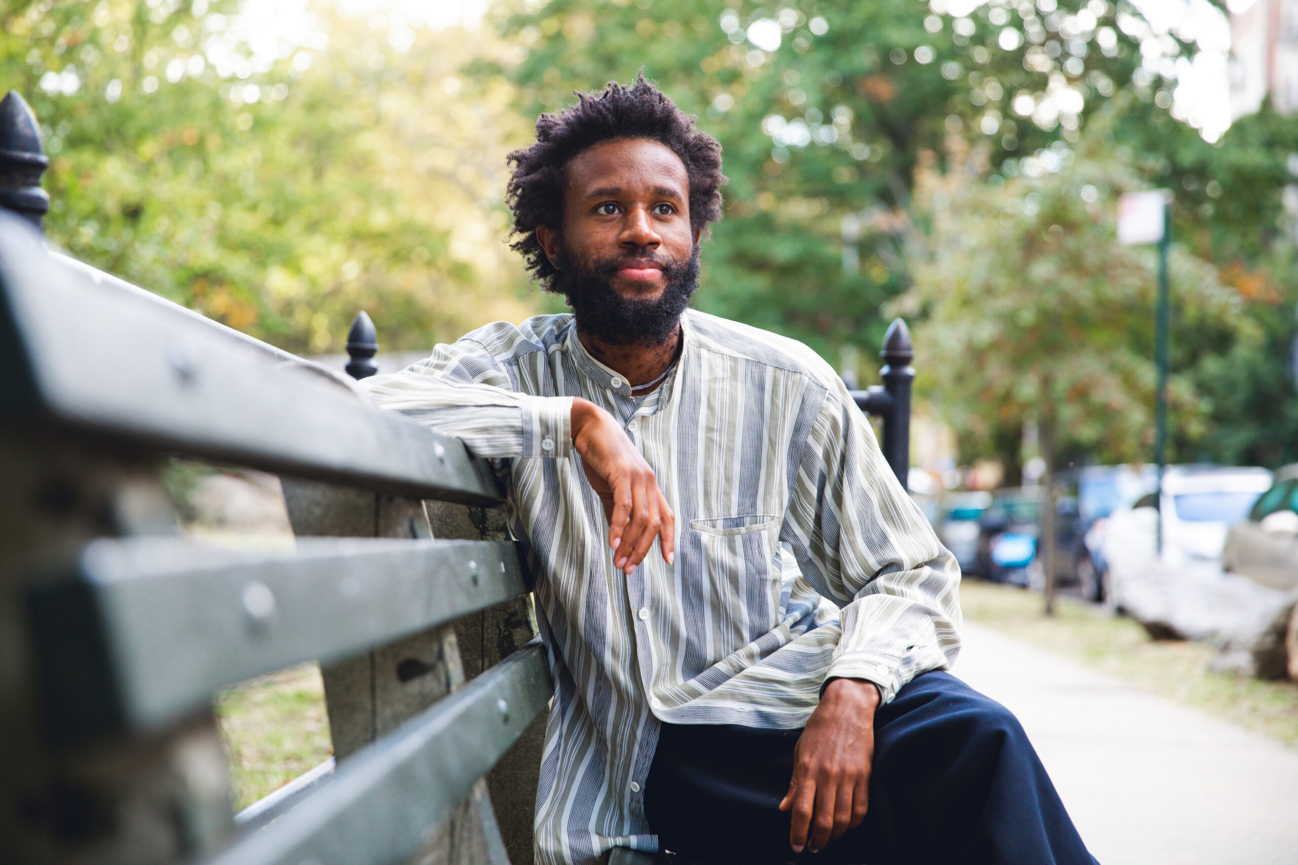 Jerome sits in a relaxed position on a park bench. One arm leans on the top of the bench, the other rests on their leg