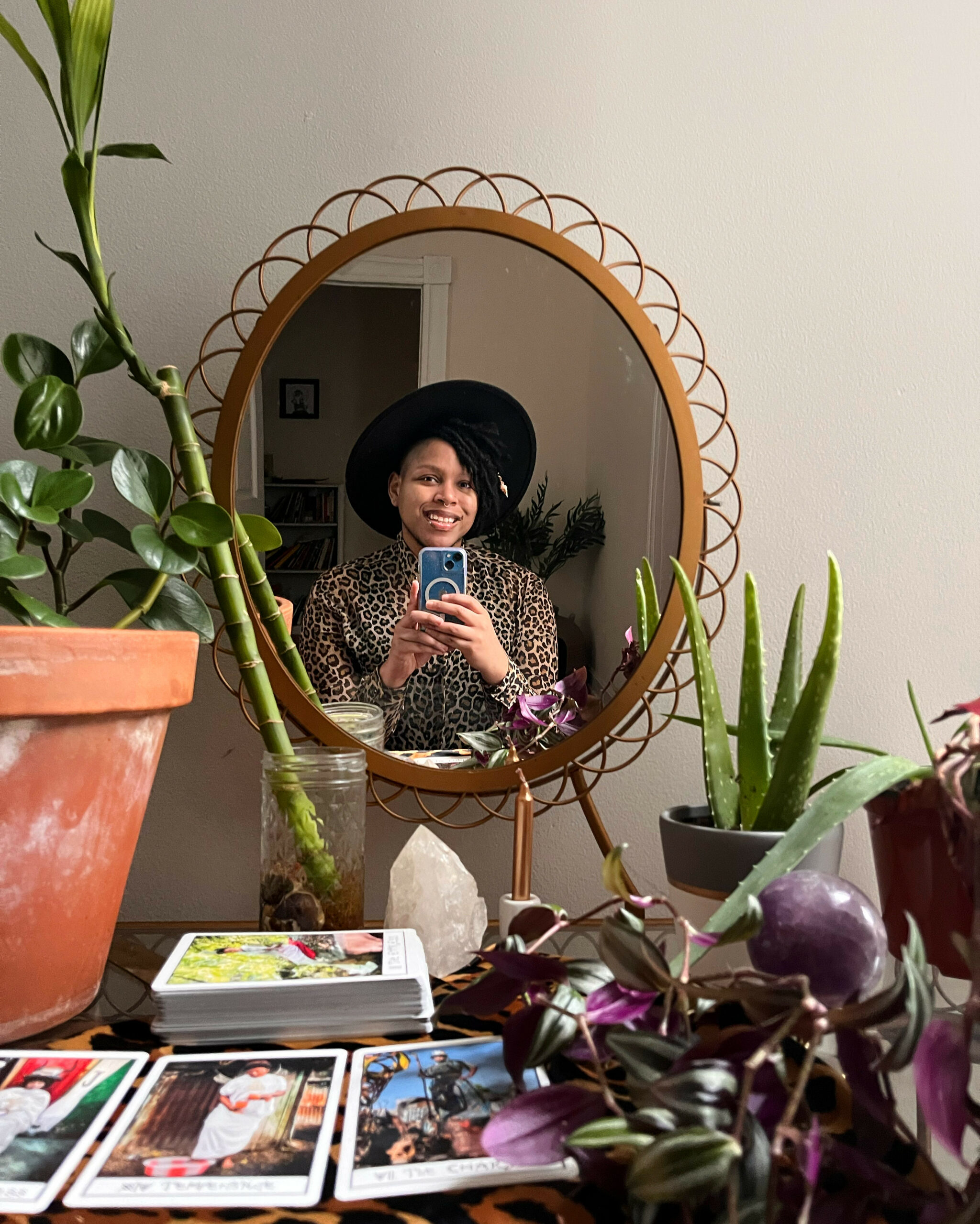 Cyrée, a black trans person, in a mirror. He’s wearing a hat and a leopard print shirt. He’s surrounded by tarot cards and plants.