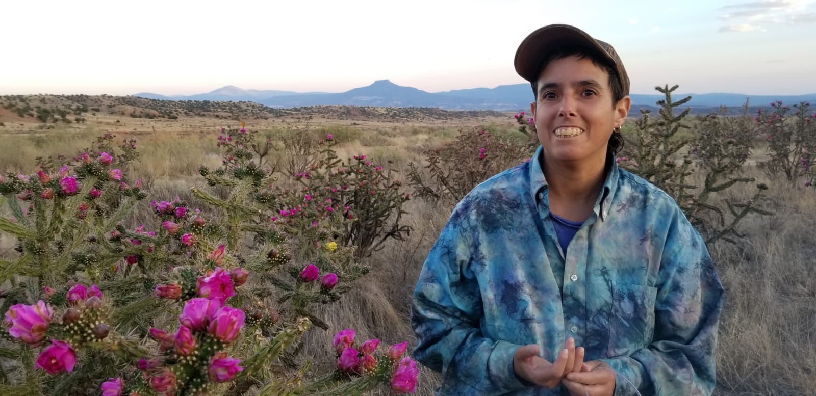 Amelia is framed to the right of the picture smiling. They are wearing a sky blue tie dye shirt and brown cap. They are pictured in Abiquiu, New Mexico, by the Chama River, surrounded by cholla cacti blooming bright pink flowers with a mountainous landscape behind.