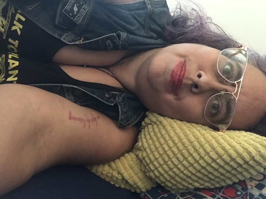 A 40ish mixed race Sri Lankan, Irish and Galician nonbinary femme with curly brown silver and purple hair, lying on a couch looking at the viewer horizontally. They have rose gold aviator frames, thick eyebrows, red lipstick and sand colored skin, and are looking at the viewer with a kind of tired but hopefully crip wonder. They wear a blue denim vest with a pin that says Neurodivergent Universe above a pink and blue image of a ringed planet, and a black tank top with yellow lettering that read Talk To Plants, Not Cops is barely visible. They have a tattoo of the words "We begin by listening" in magenta cursive script on their left arm.