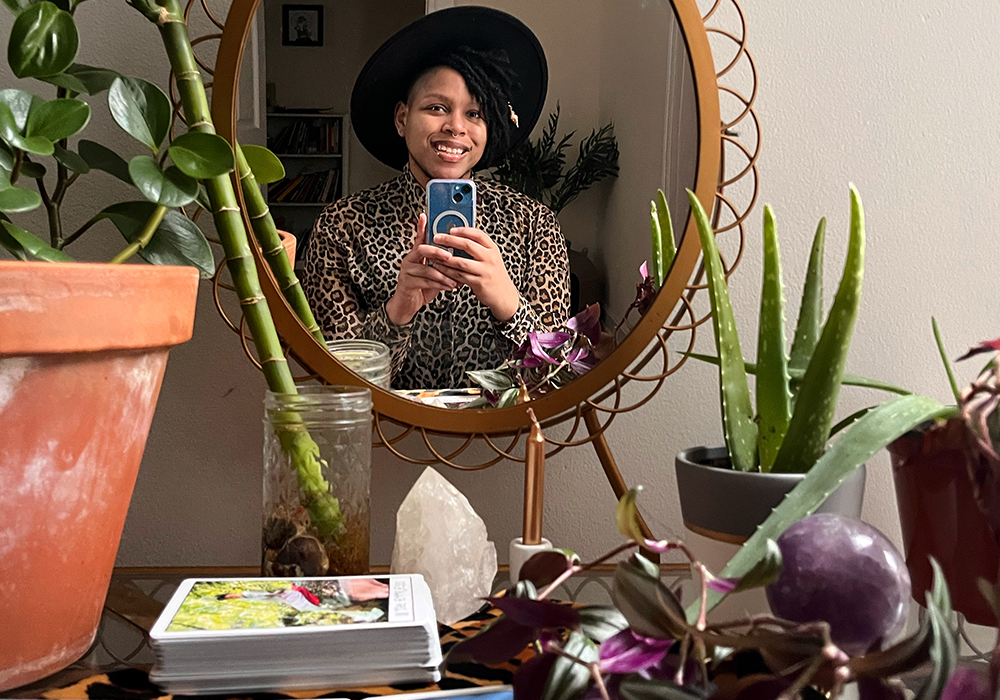Cyrée, a black trans person, in a mirror. He’s wearing a hat and a leopard print shirt. He’s surrounded by tarot cards and plants.