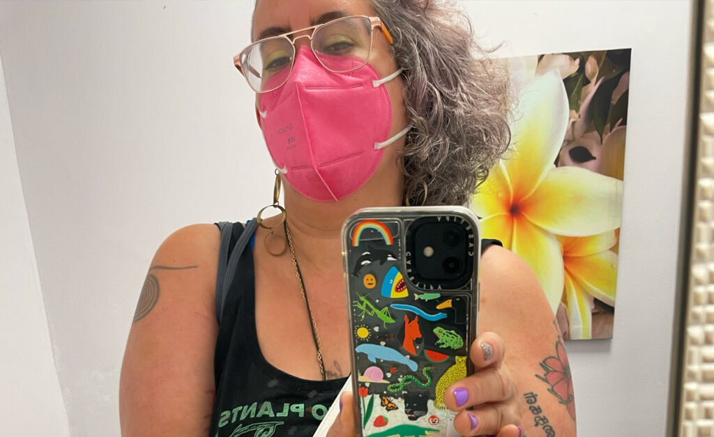 a Sri Lankan, Irish and Galician nonbinary femme with curly brown silver and purple hair and an undercut, wearing a “talk to plants, not cops” navy and turquoise crop top, tiny black denim shorts and a hot pink KN95 mask, takes a mirror selfie with a phone with glitter and many kinda of brightly colored animals and plants on it.