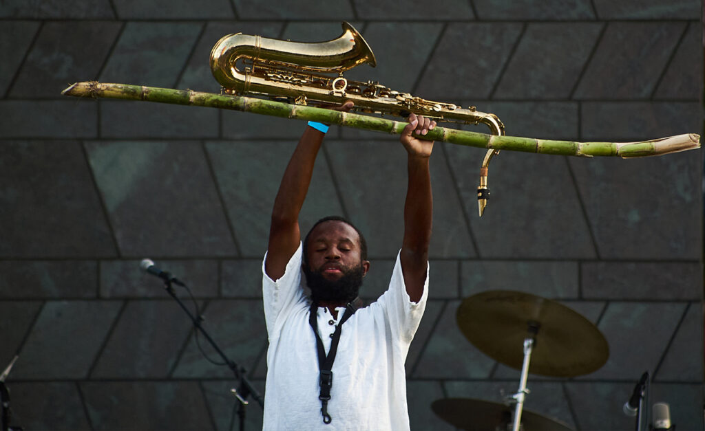 Jerome in a white t-shirt holds aloft a saxophone and long stem of sugar cane above their head