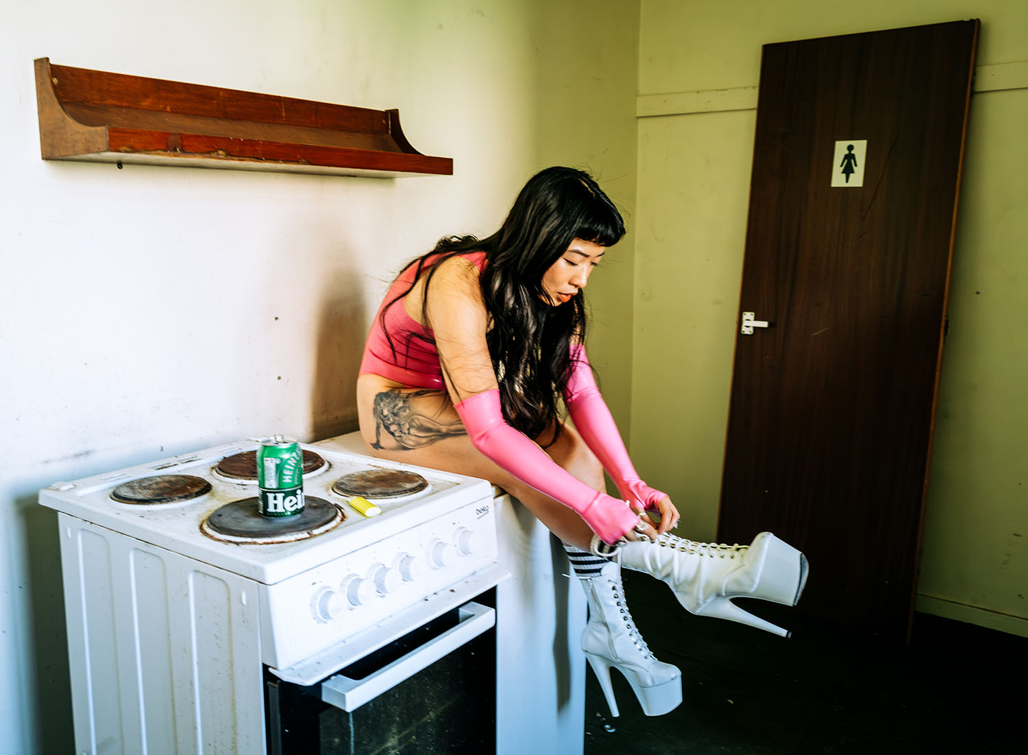 Chao-Ying Rao, an East Asian woman with long dark hair. She wears a high heeled white boots, pink latex singlet and elbow length gloves. She is sat on top of a fridge in a kitchen like space.