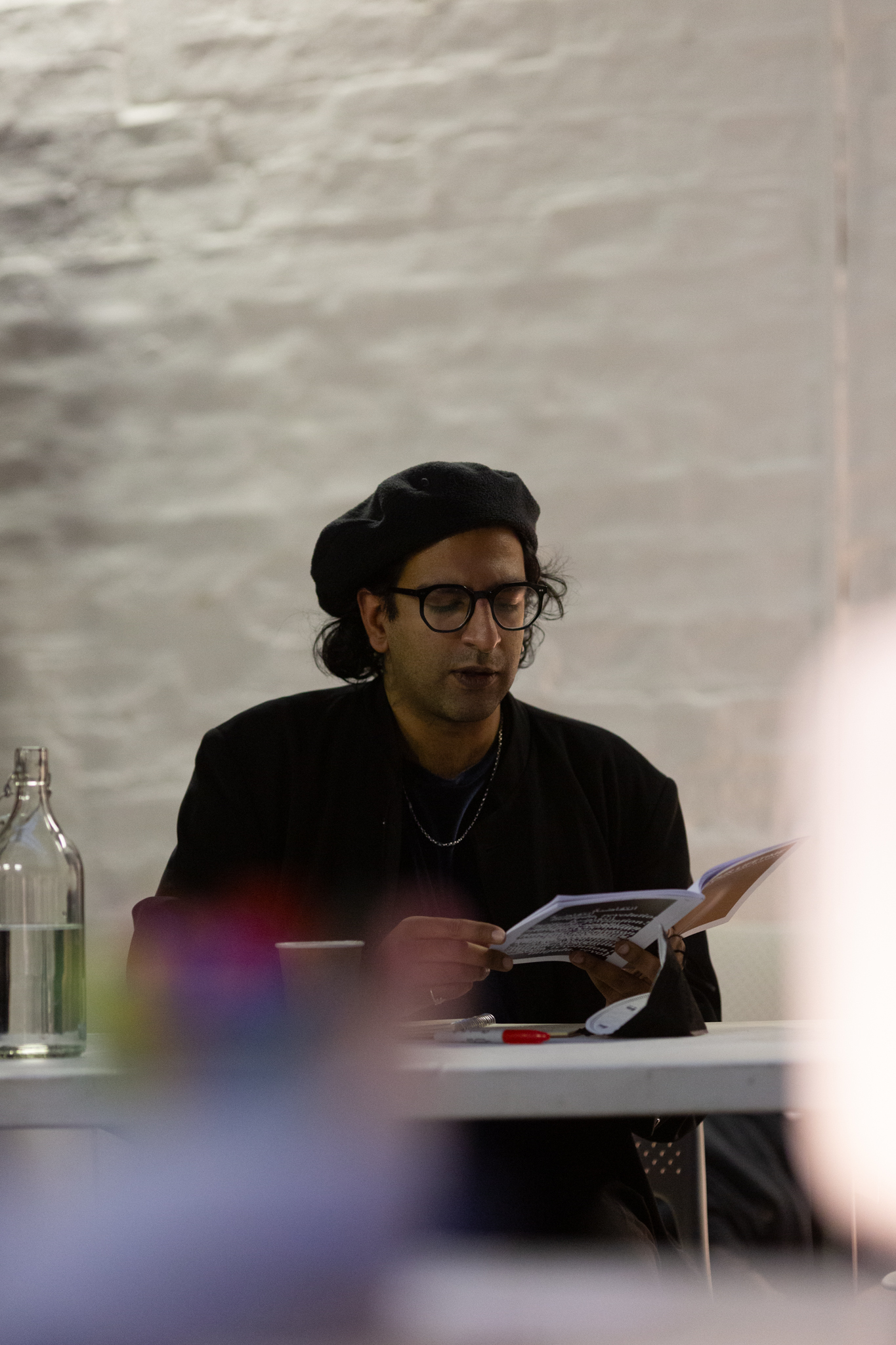 Hussein Mitha sits at a table reading from their workbook "In Our Lifetime: A anti-imperialist resource"