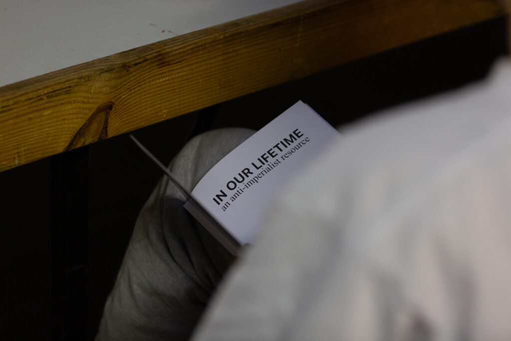 An open booklet rests on someone's lap, we see the title "IN OUR LIFETIME an anti-imperialist resource"