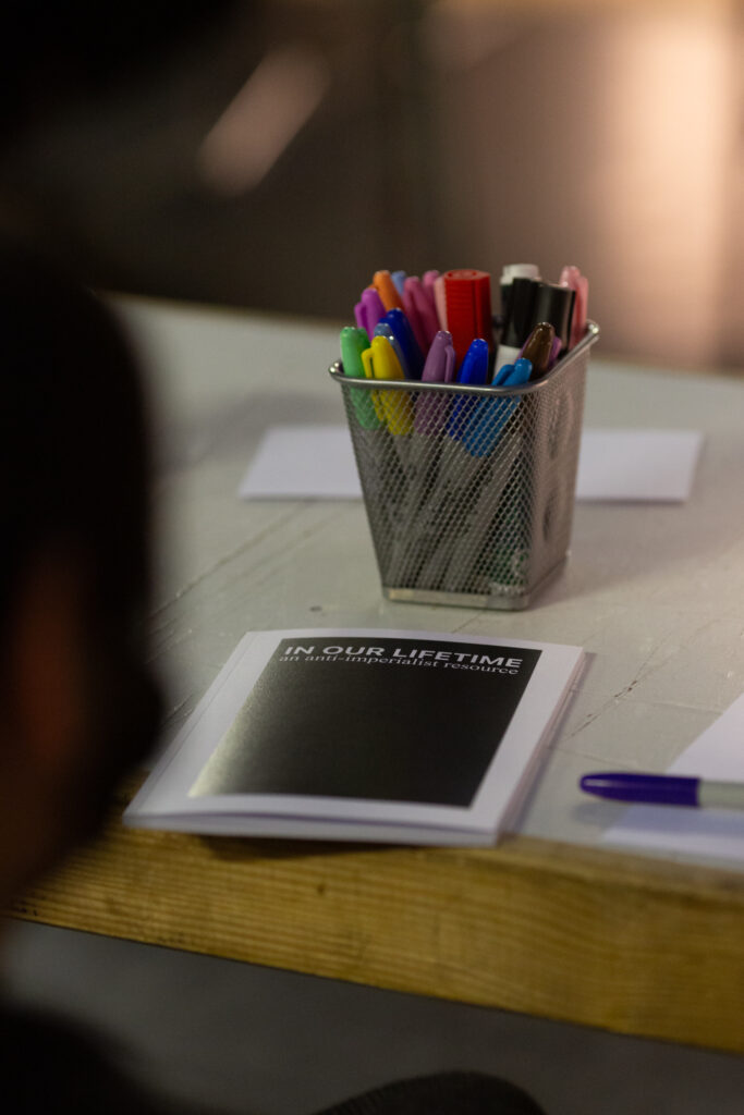 A table with a book titled "IN OUR LIFETIME: an anti-imperialist Resource", new to is a small metal basket holding lost of maker pens of differing colours and bits of paper.