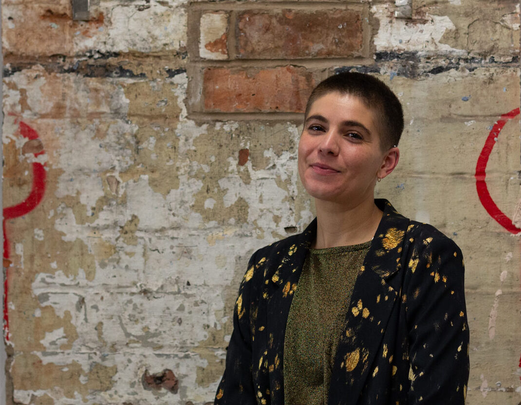 Portrait of Emilia Beatriz, They stand smiling infront of a brick wall wearing a black and gold outfit