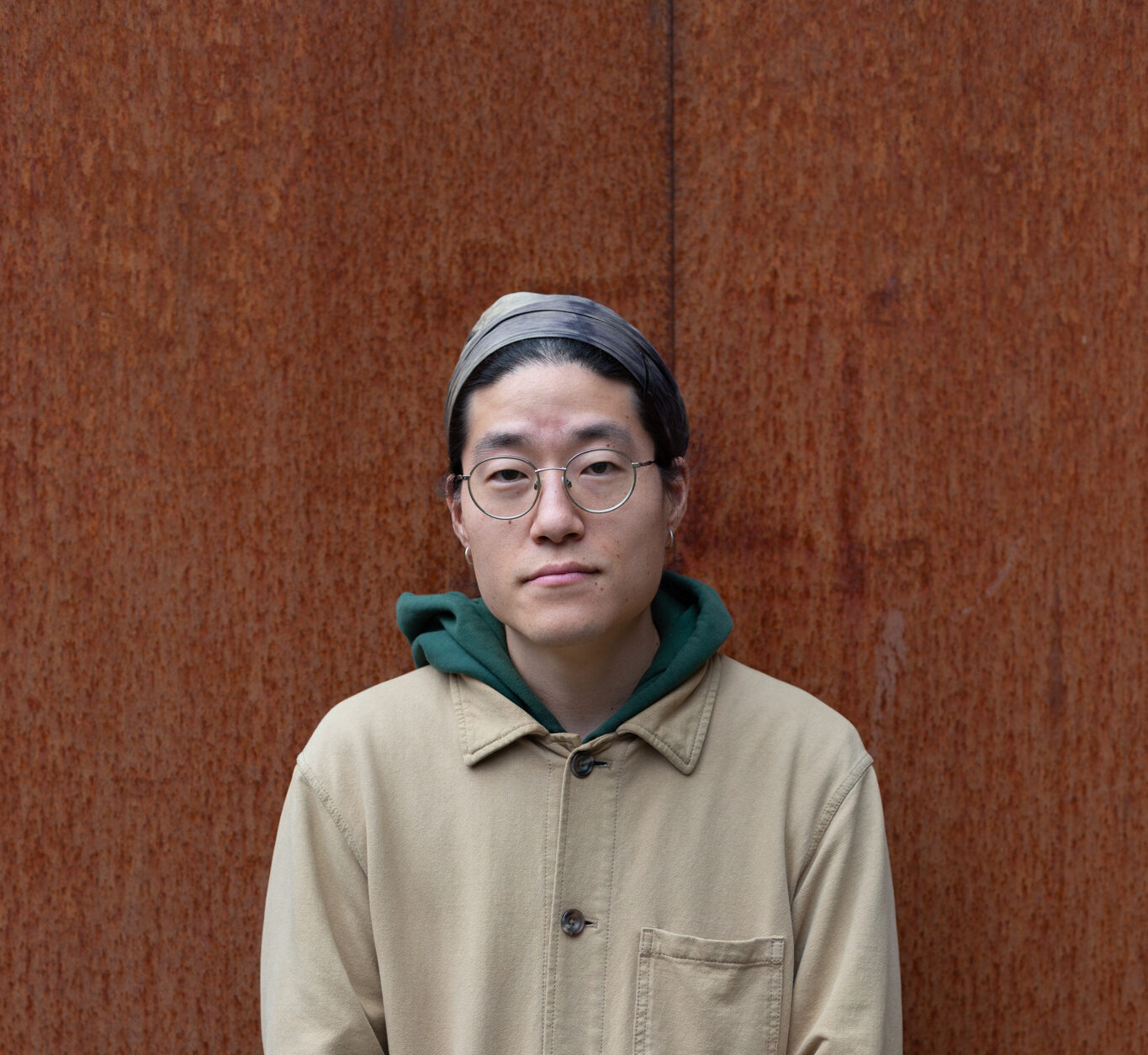 Sunik Kim stands against a rusted wall. They are wearing round rimmed glasses, a grey head scarf and a green hoodie under a beige button up shirt