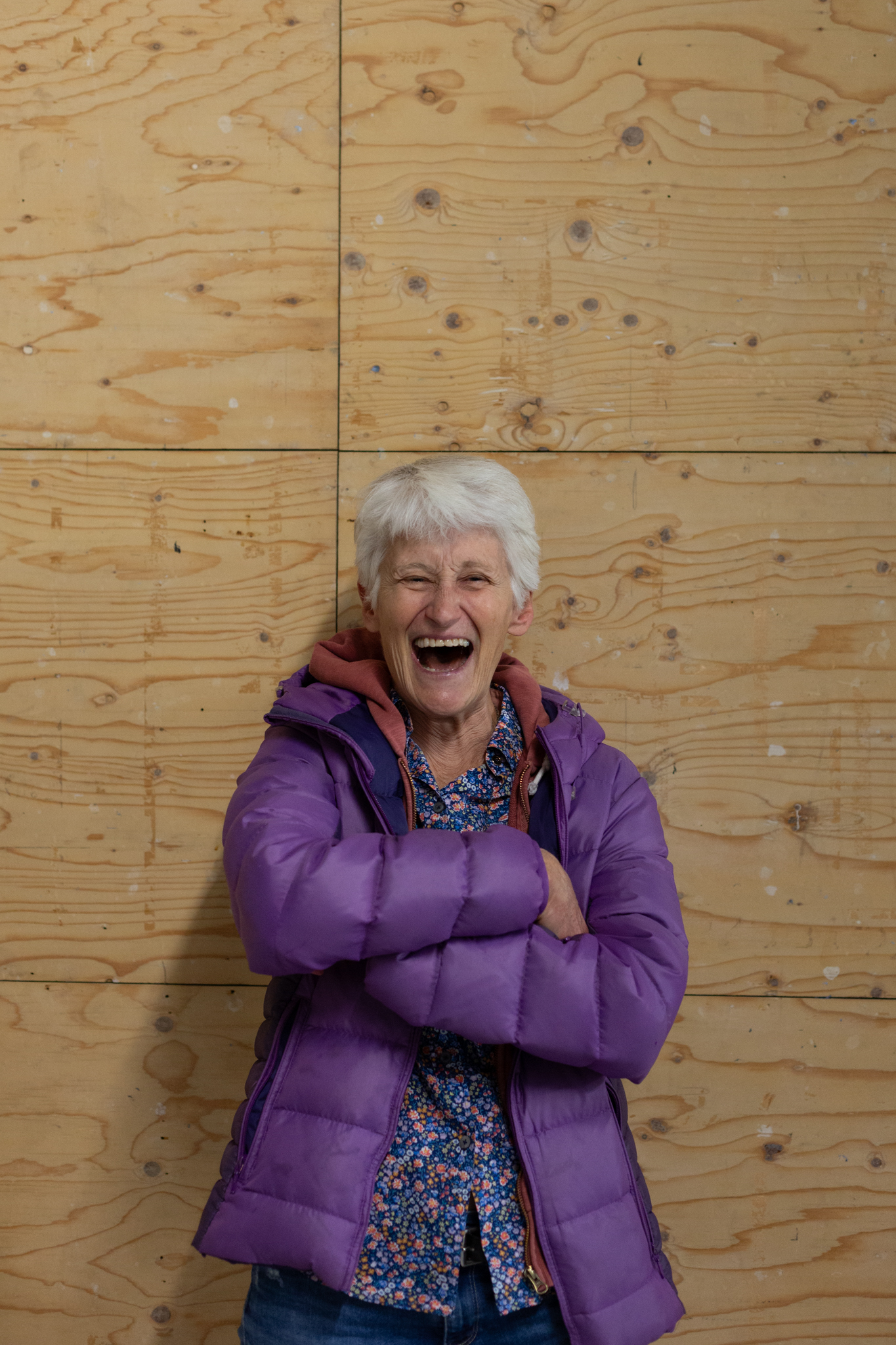 Elizabeth A. Povinelli smiles cheerfully with a wide open mouth. They have short grey hear and wear a bright purple padded jacket over a floral blue shirt