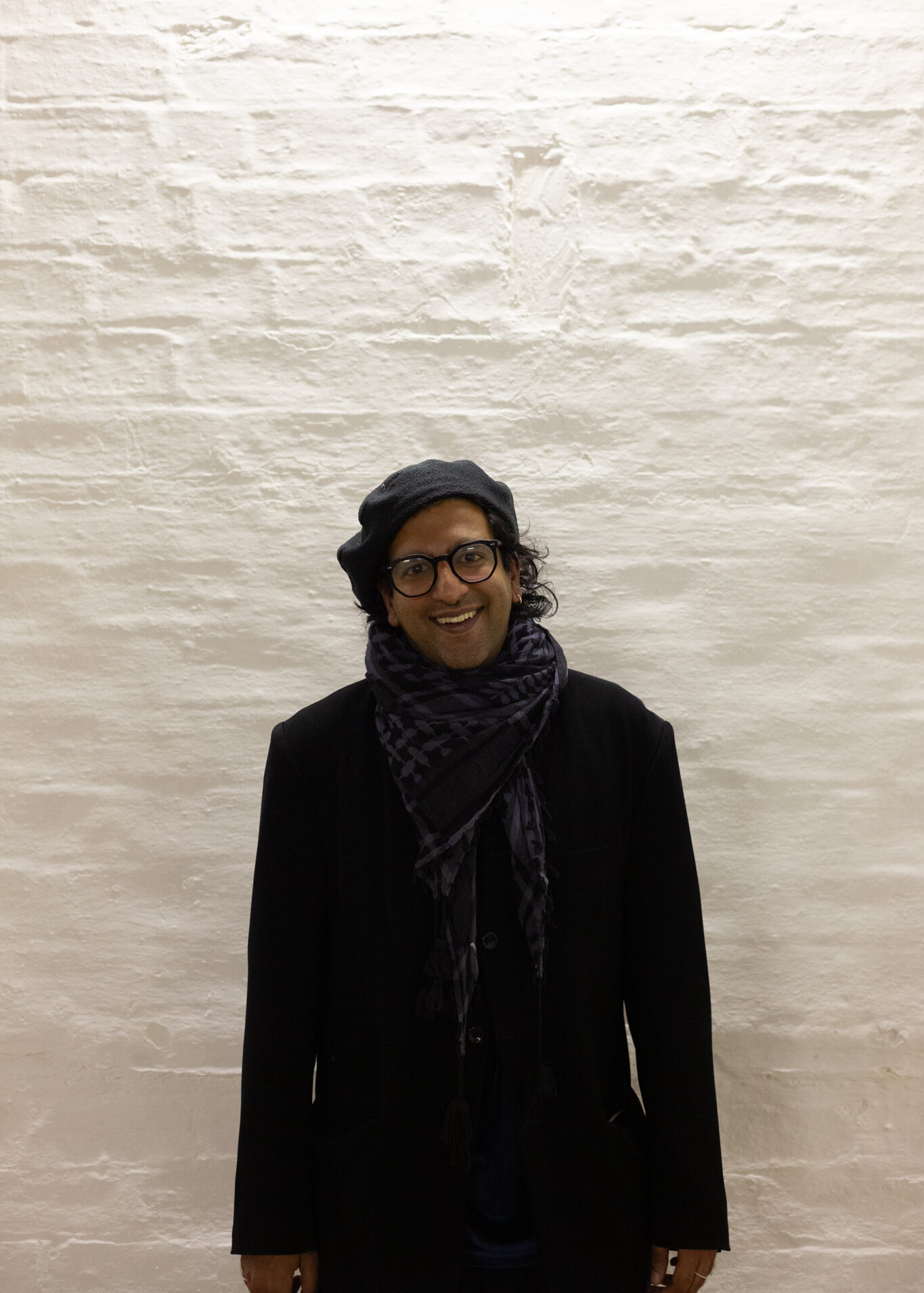 Hussein Mitha stands smiling. They wear dark rimmed glasses, a grey berry & keffiyeh scarf