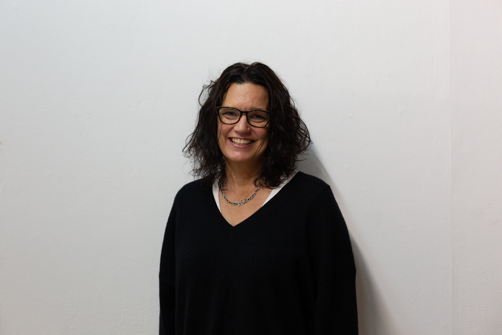Leanne Betasamosake Simpson stands smiling broadly. They have dark shoulder length wavy hard and dark rimmed glasses