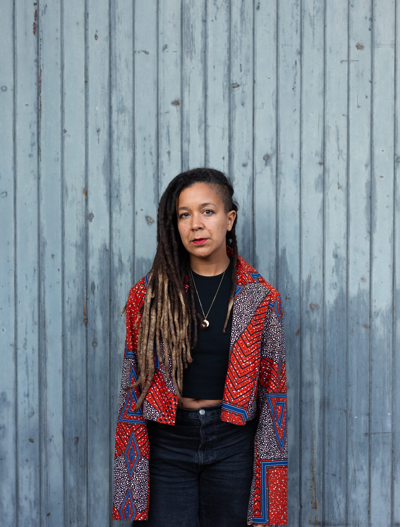 Robyn Maynard stands against a blue-grey wood clad wall. They have long braided and wear a red patterned jacket