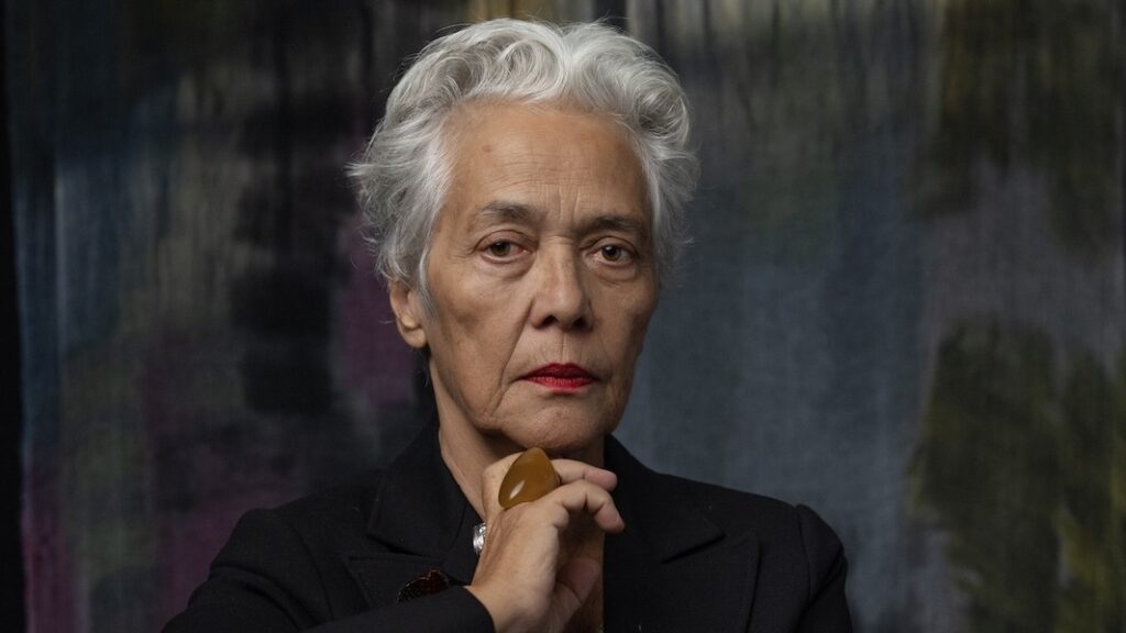An older woman with olive-brown skin, short white and grey hair, and brown eyes. She is posed resting her chin on the knuckle of her hand thoughtfully. She wears a black blazer, chunky colourful jewellery, and minimalist makeup. She's posed in front of a large abstract painterly background.