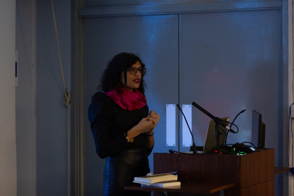 Nat Raha stands at a lectern talking into a mic and is addressing an audience. She is wearing a bright pink scarf contrasting a dark outfit.