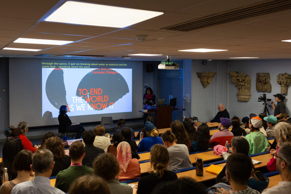 Leanne Betasamosake Simpsons stands infront of a busy lecture theatre filled with people. Behind Leanne is projected image saying "Episode 11: To End the World As We Know It.
