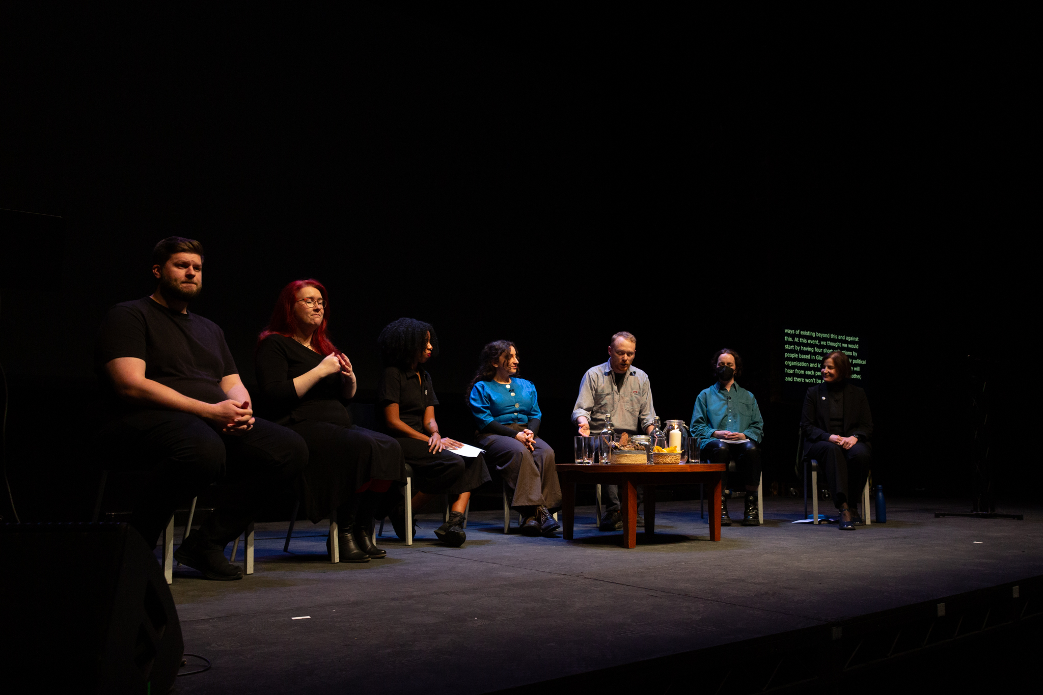 A group of people are seated on a spotlit stage in a dissussion style event.