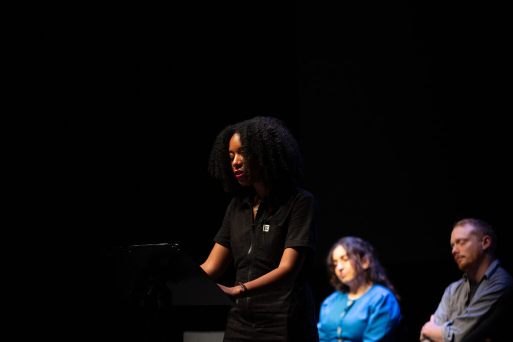 Gracie Mae Bradley stands on stage in front of a lectern delivering their performed reading.