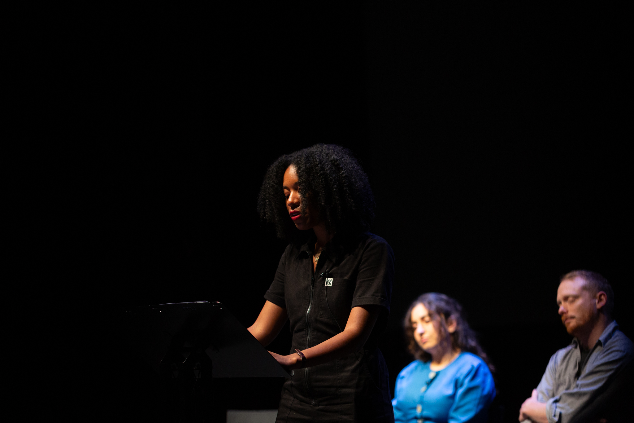 Gracie Mae Bradley stands on stage in front of a lectern delivering their performed reading.
