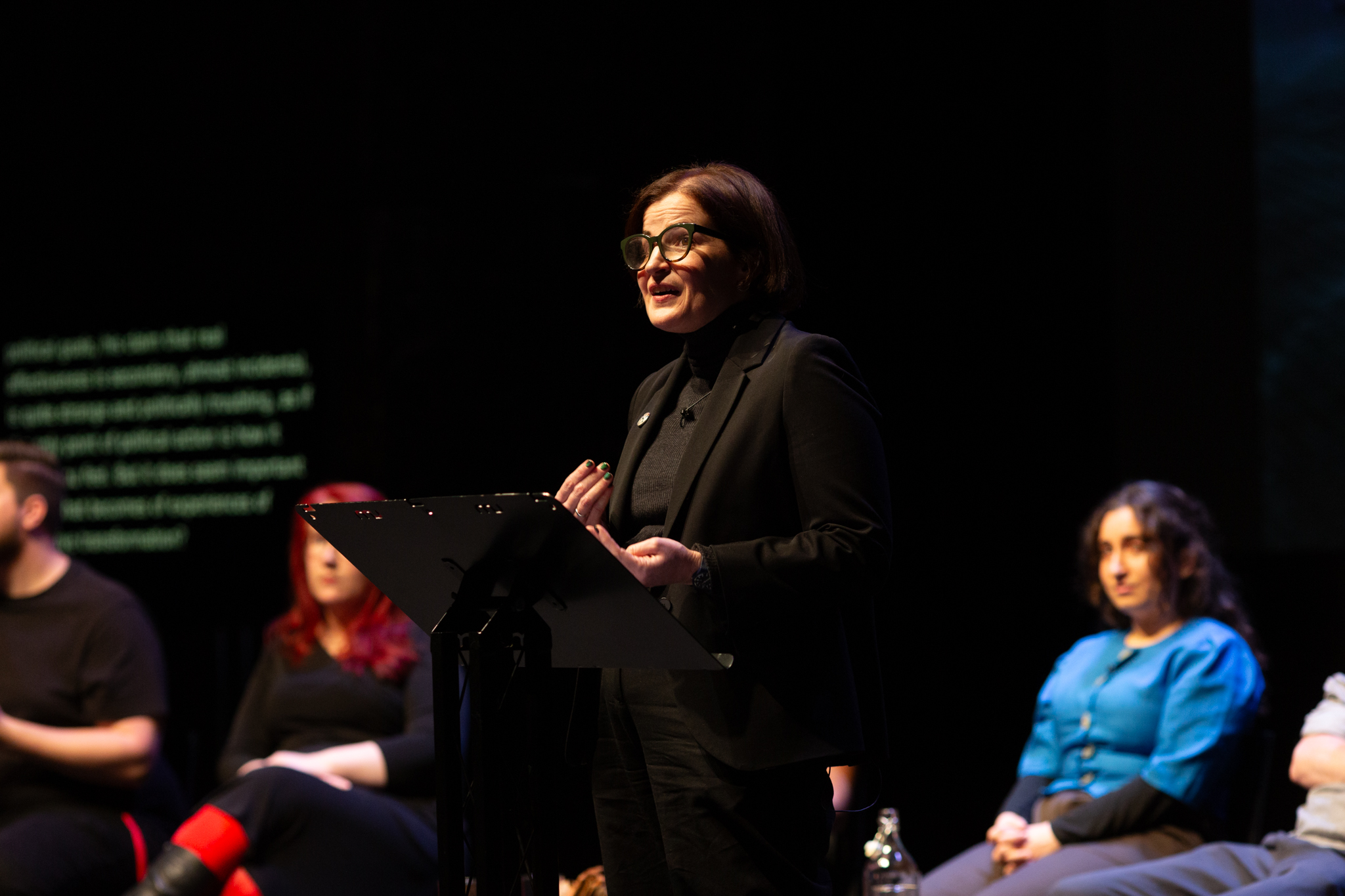 Hannah Proctor stands on stage in front of a lectern delivering their performed reading.