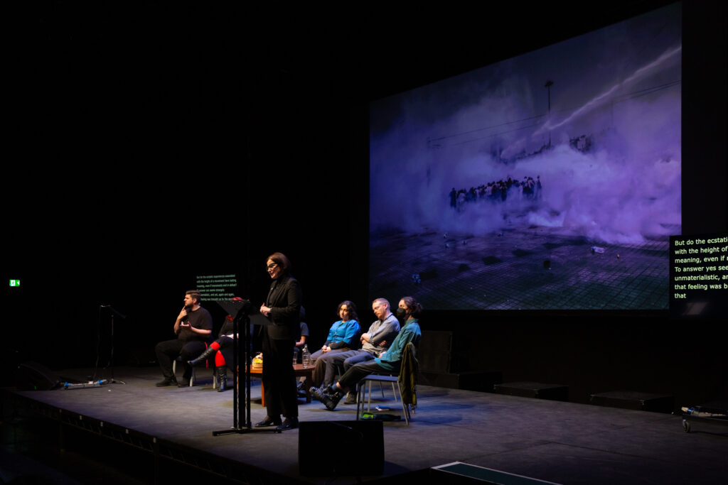 Hannah Proctor stands on stage in front of a lectern delivering their performed reading. Behind Hannah is a project image of a crowd in a smoke filled city street, giving the impression of a conflict or war environment.
