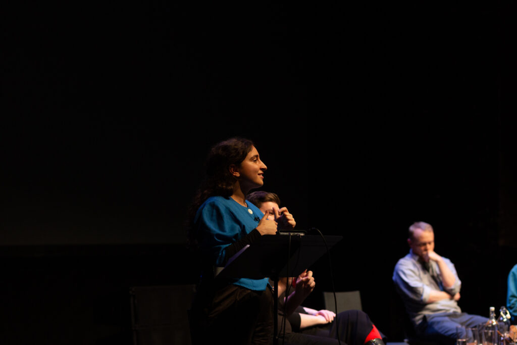 Masa Nazzal stands at a lectern delivering their performed reading to an audience.