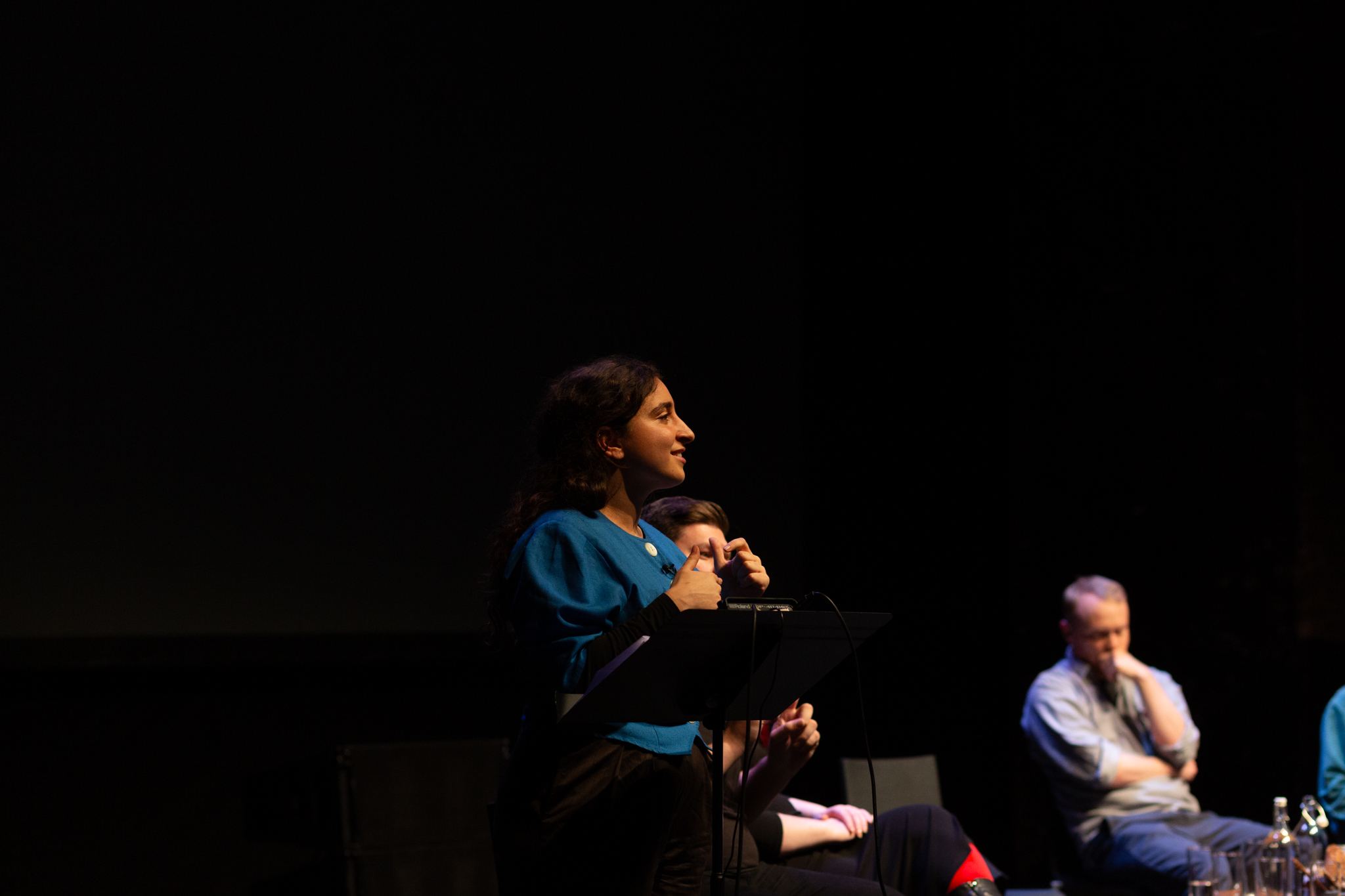 Masa Nazzal stands at a lectern delivering their performed reading to an audience.