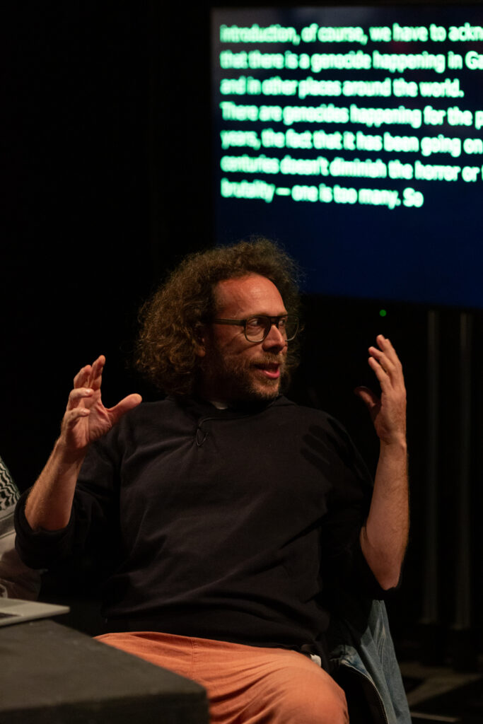 Amilcar Packer talks expressively to an audience, gesturing with their hands. Being them is a screen with text from live captioning