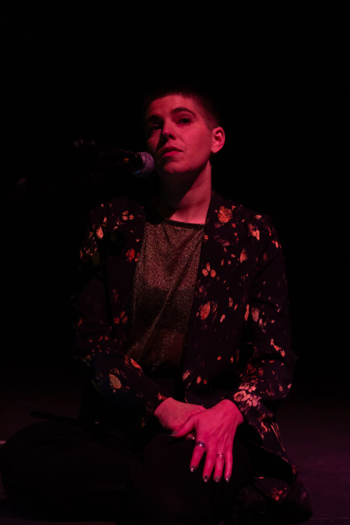 Emilia Beatriz is seated speaking into a microphone. They are cast in a reddish light stage lights,