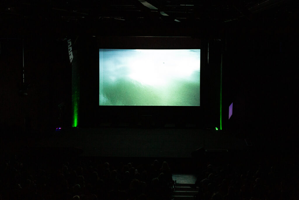 Green lights glow upwards against a wall at either side of glowing projected screen which features green shimmering water.