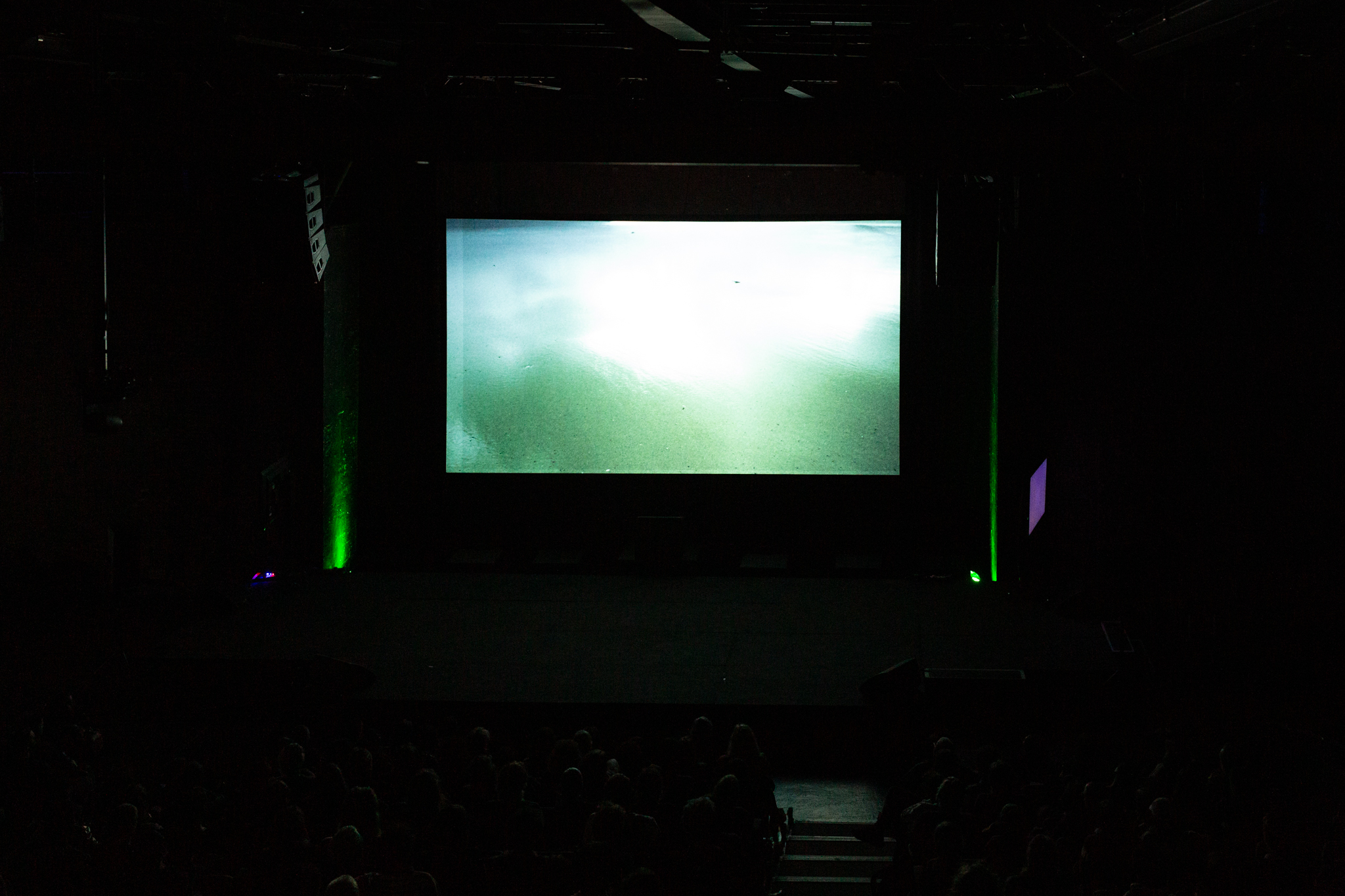 Green lights glow upwards against a wall at either side of glowing projected screen which features green shimmering water.