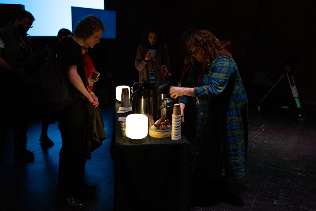 Someone stands behind a table serving Grief Tea from a flask to audience members at Emilia Beatriz's screening of "BARRUNTO"