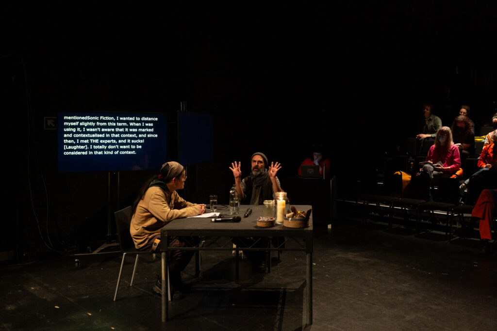 Rashad Becker in conversation with Sunik Kim. Rashad talks expressively the palm of his hands face outwards and are lifted close to his face.
