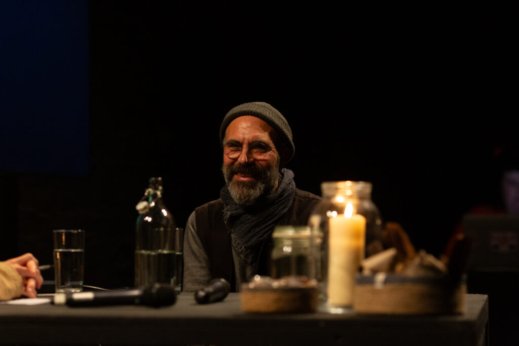 Rashad is smiling. He is sat at a black table which has two microphones & a lit candle in a glass jar