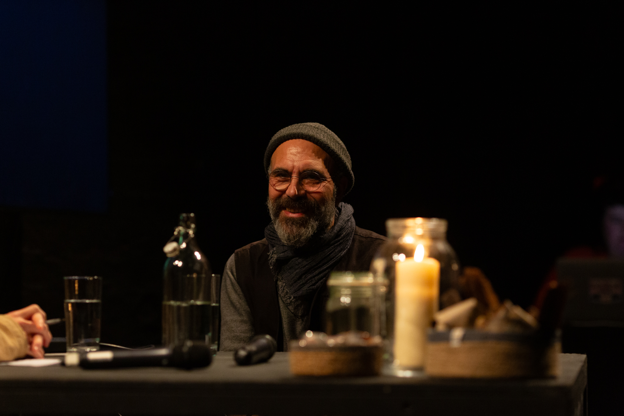 Rashad is smiling. He is sat at a black table which has two microphones & a lit candle in a glass jar