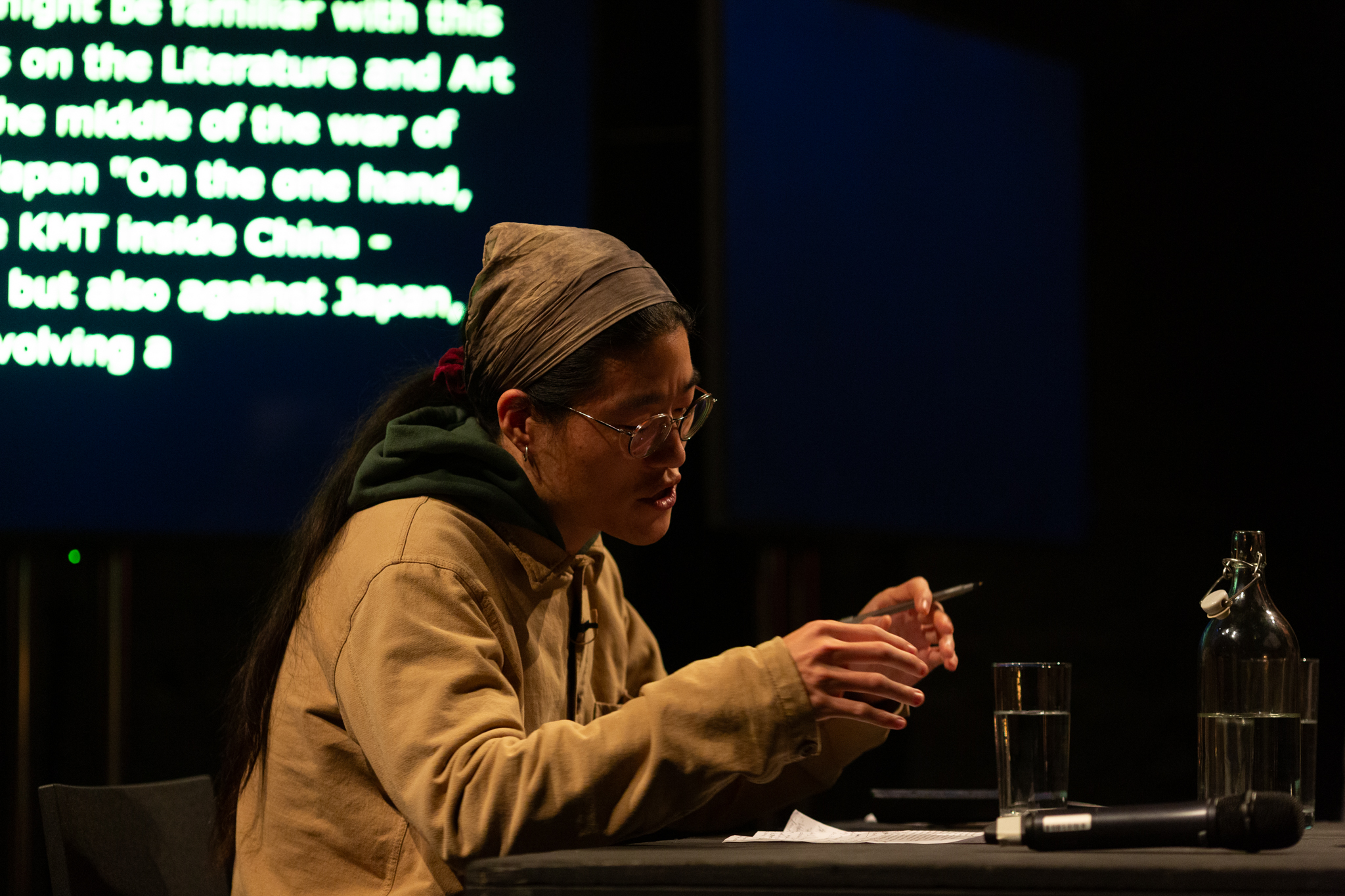 Sunik Kim talks expressively with gestured hands. They look down a notes on a table which they are referring to and taking notes.