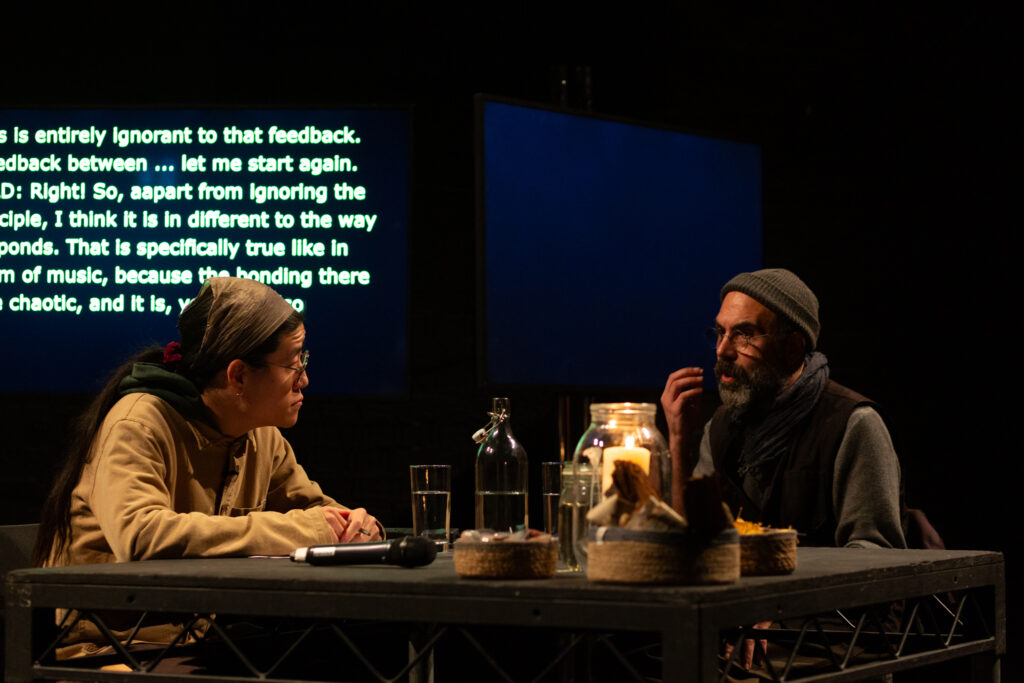 Rashad Becker & Sunik Kim are in conversation. Rashad is talking and Sunik is listening intently. They are sat a square black table which has a lit candle in a glass jar and small baskets, glasses filled with water and a microphone.