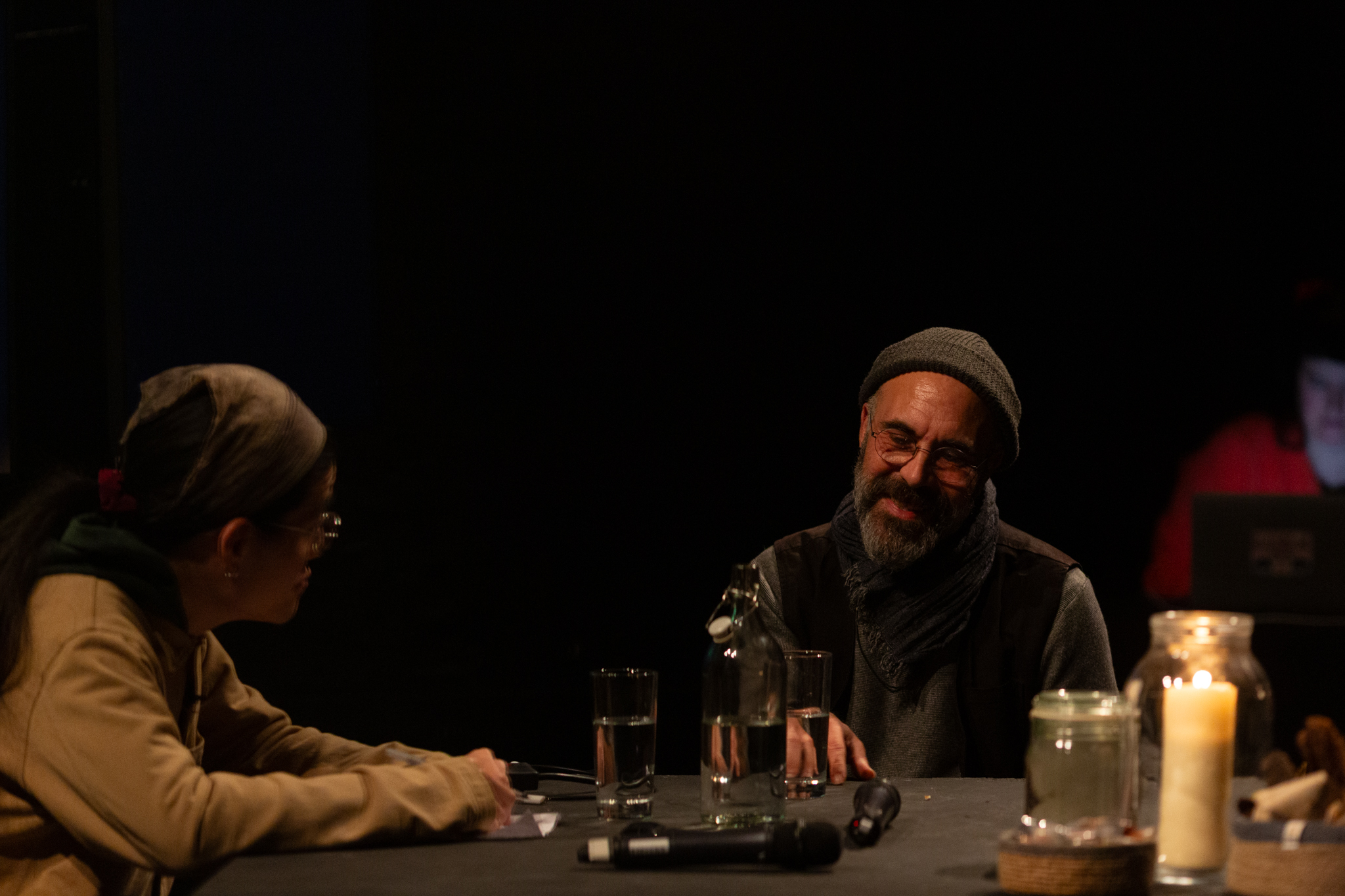 Rashad Becker and Sunik Kim are in conversation together. They are sat at a black table which has two microphones, water glasses and a lit candle in a glass jar.