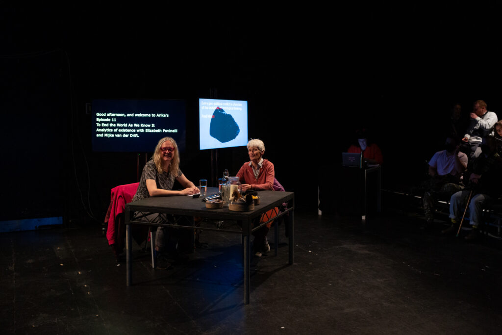 Elizabeth A. Povinelli and Mijke van der Drift sit at black table in a black box theatre. There is audience to the right of the image and behind Mijke & Elizabeth are two screens for live captioning.