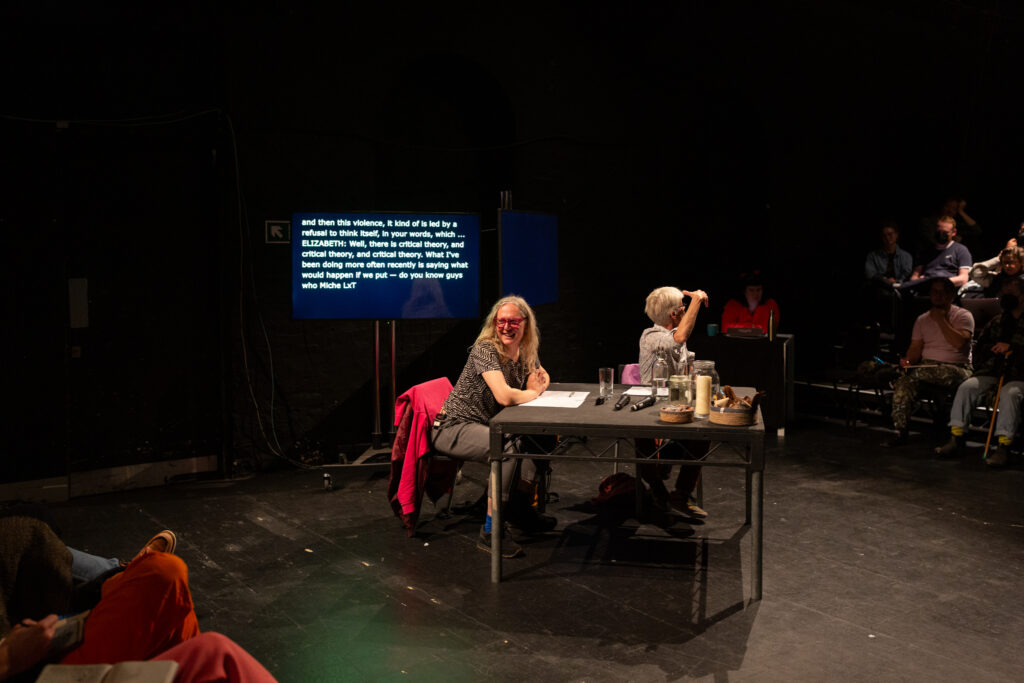 Elizabeth A. Povinelli and Mijke van der Drift sit at black table in a black box theatre. There are audience is seating banks at either side of them. Mijke smiles to the audience on their right and Elizabeth looks at audience to their left while gesturing expressively