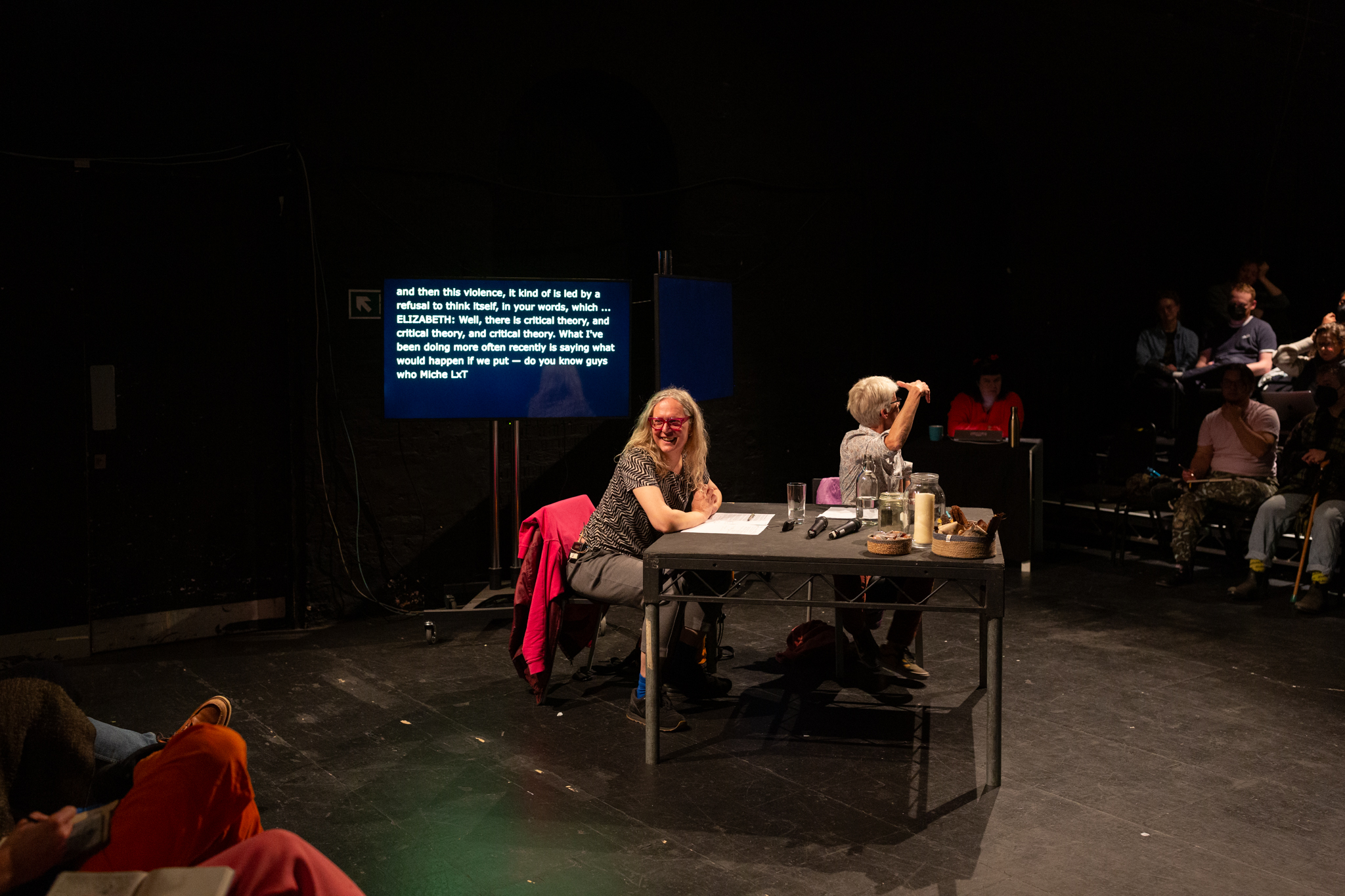 Elizabeth A. Povinelli and Mijke van der Drift sit at black table in a black box theatre. There are audience is seating banks at either side of them. Mijke smiles to the audience on their right and Elizabeth looks at audience to their left while gesturing expressively