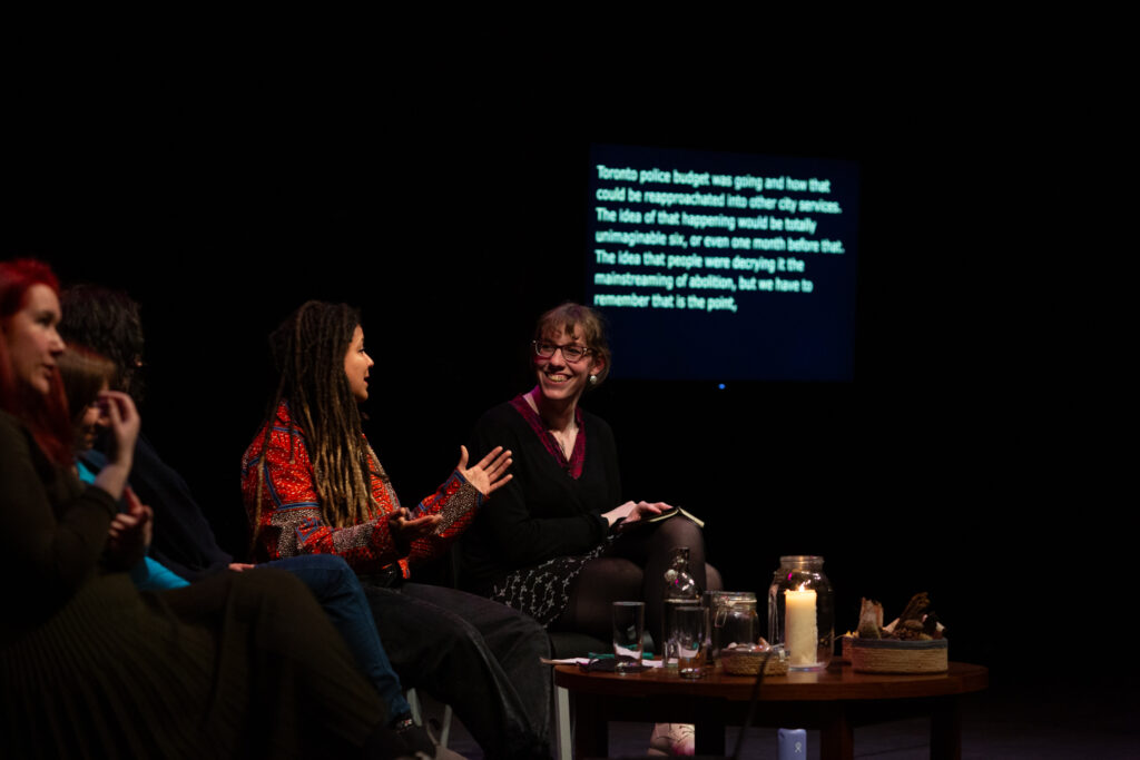 Harry Josephine Giles smiles at Robyn Maynard who is talking and expressively gesturing with their hands. Behind them in a screen with blurred text.