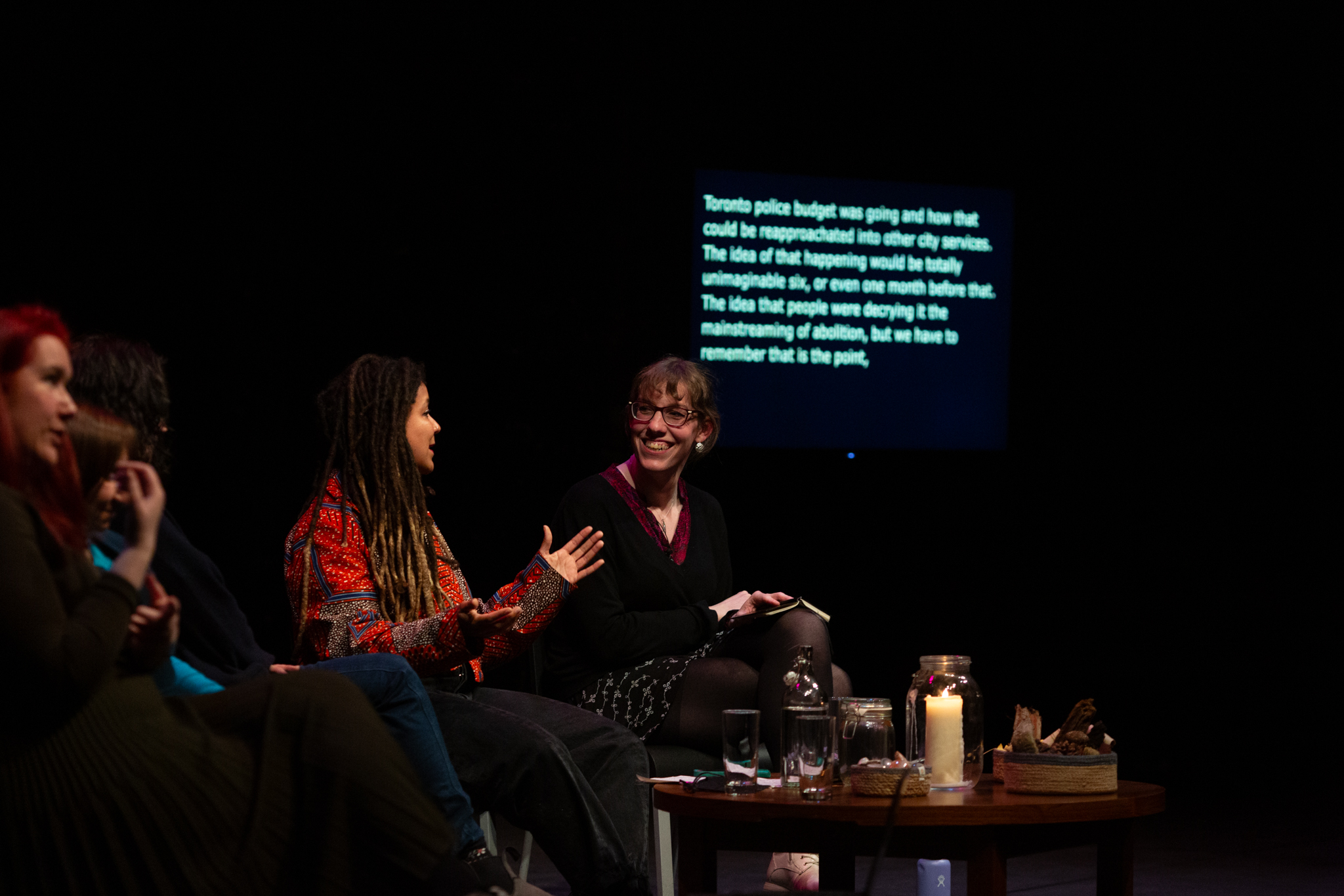 Harry Josephine Giles smiles at Robyn Maynard who is talking and expressively gesturing with their hands. Behind them in a screen with blurred text.
