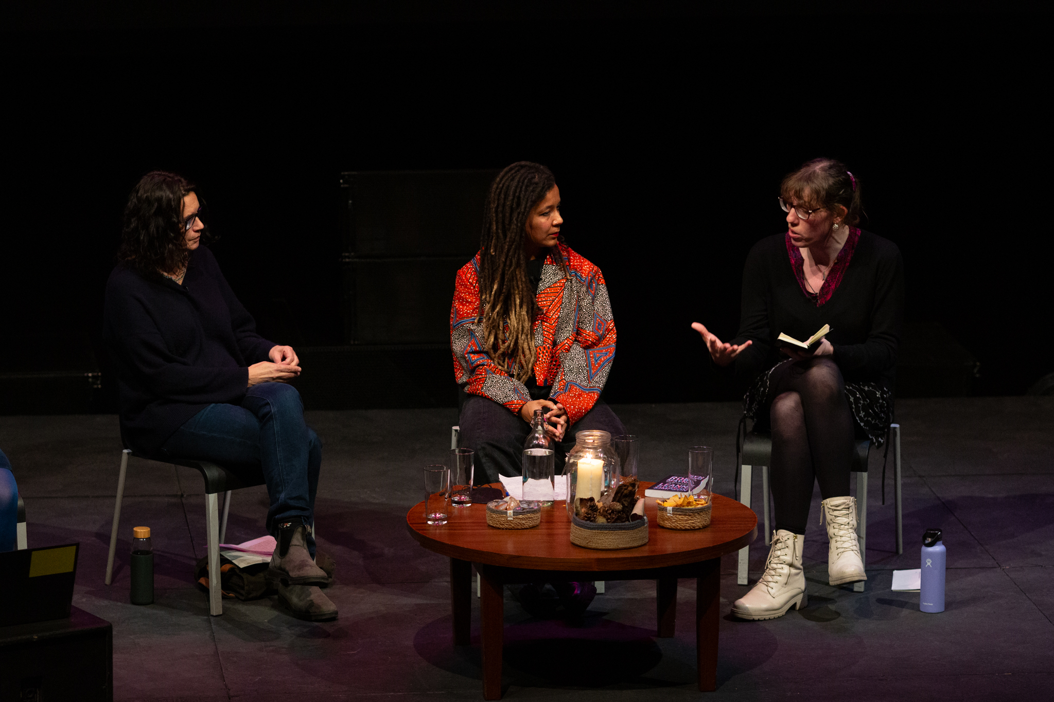Leanne Betasamosake Simpson, Robyn Maynard & Harry Josephine Giles sit in discussion on stage. Harry Josephine Giles is speaking with a notebook in hand. A small table with candles, drinks, and snacks is between them. The setting is intimate and dimly lit.