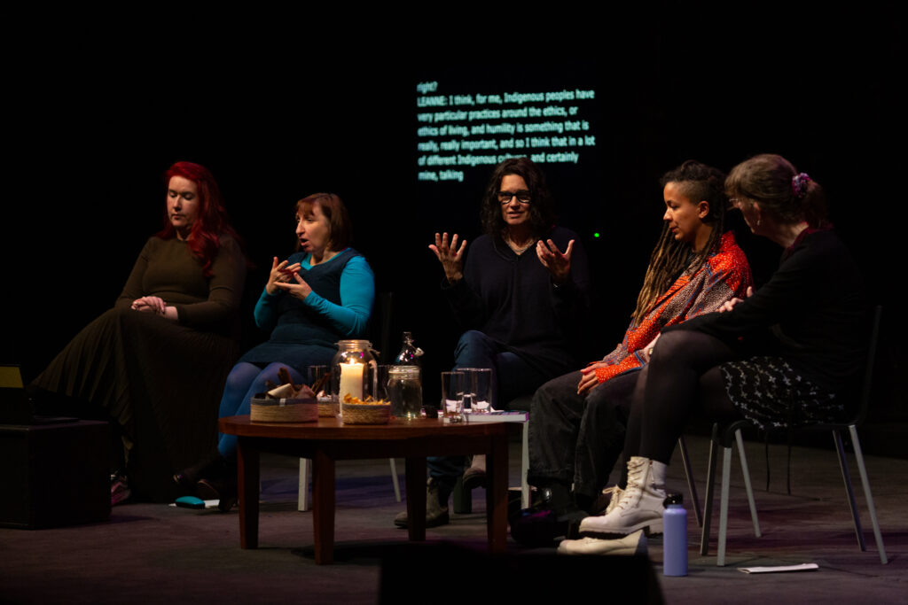 Leanne Betasamosake Simpson, Robyn Maynard & Harry Josephine Giles sit in discussion on stage. Leanne Betasamosake Simpson is speaking and gesturing with open hands. To the right sit two sign language interpreters. A small table with candles, and drinks is between them. The setting is intimate and dimly lit.