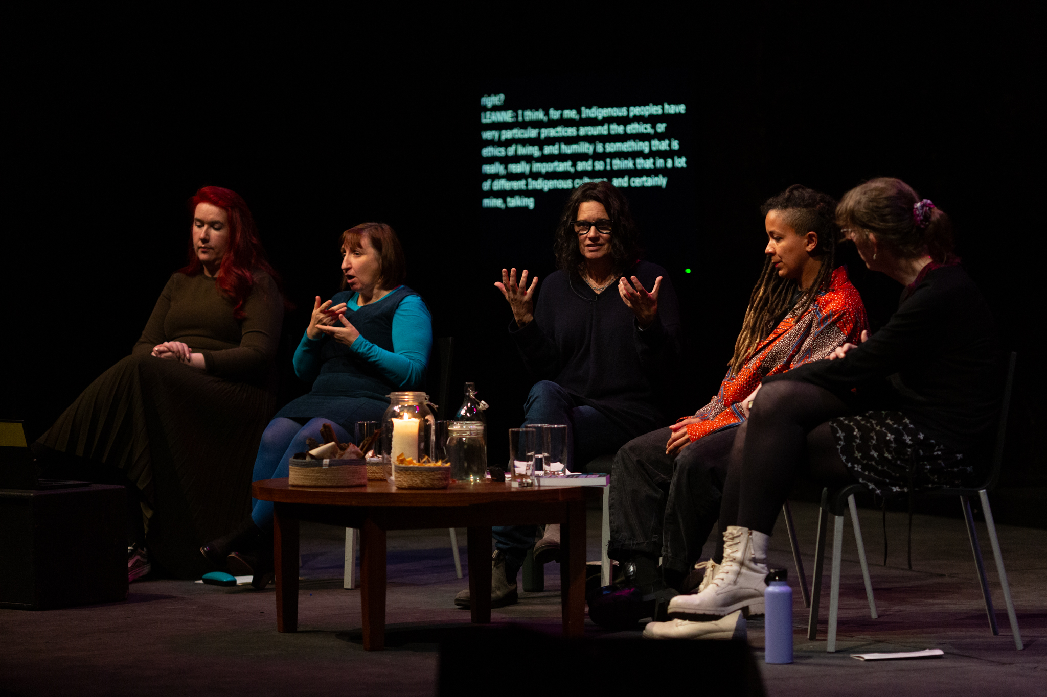 Leanne Betasamosake Simpson, Robyn Maynard & Harry Josephine Giles sit in discussion on stage. Leanne Betasamosake Simpson is speaking and gesturing with open hands. To the right sit two sign language interpreters. A small table with candles, and drinks is between them. The setting is intimate and dimly lit.