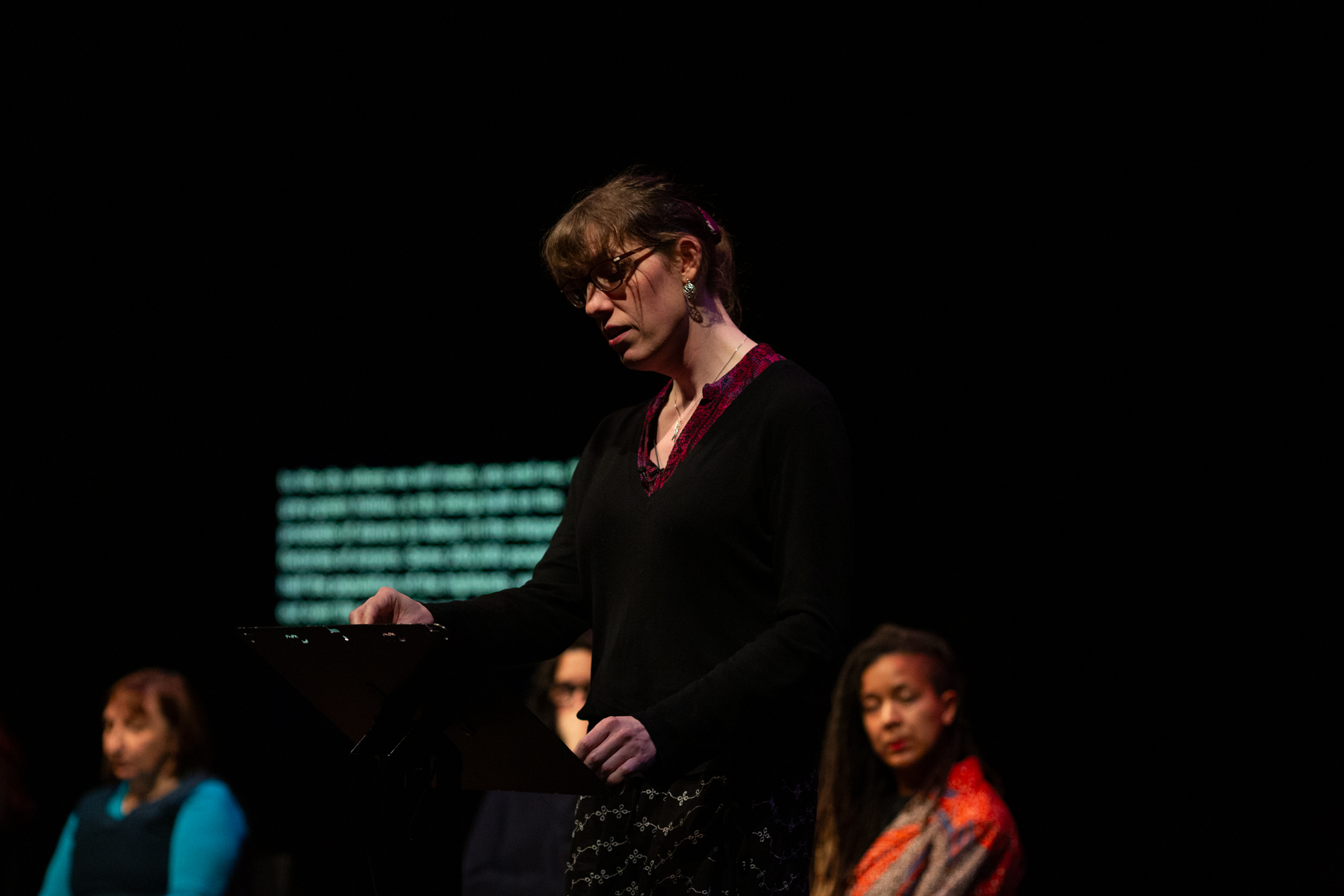 Harry Josephine Giles stands at a lectern performing a reading, she wears a black outfit with a flash of red around the collar, earring and necklace.
