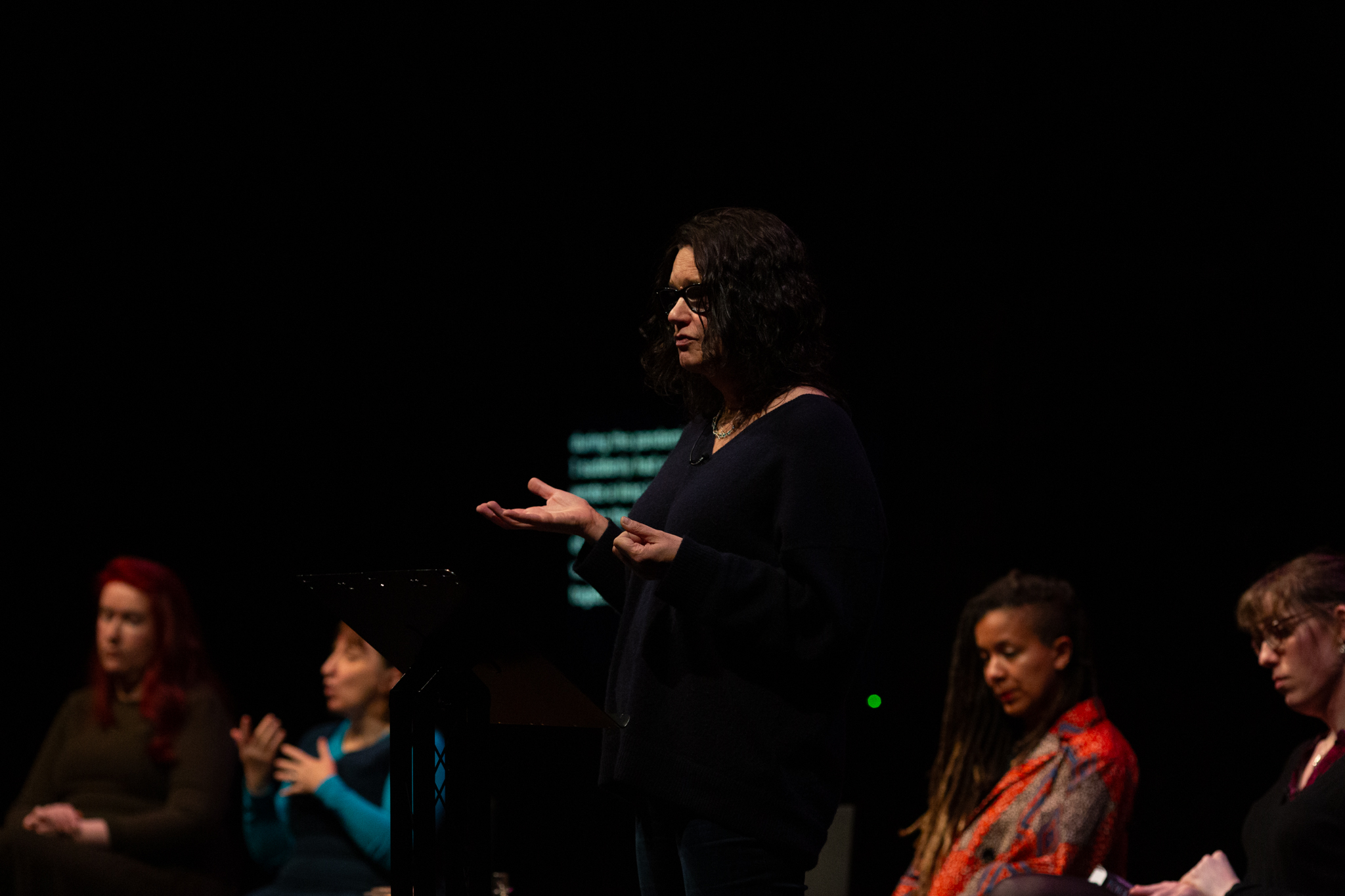Leanne Betasamosake Simpson speaks at a podium with open hands, while four others sit behind her. The background is dark, with a faint screen showing text.